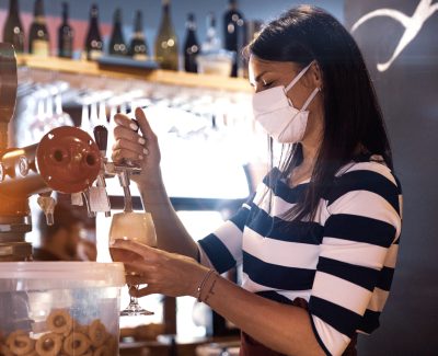 Bartender serves a fresh beer in a pub in the pandemic days, wearing protective face mask.